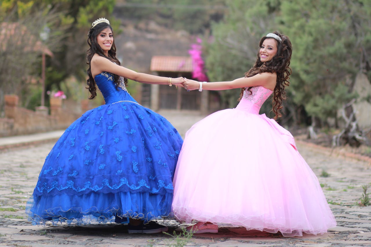 Services Two young women in elegant gowns holding hands outdoors, smiling at the camera.